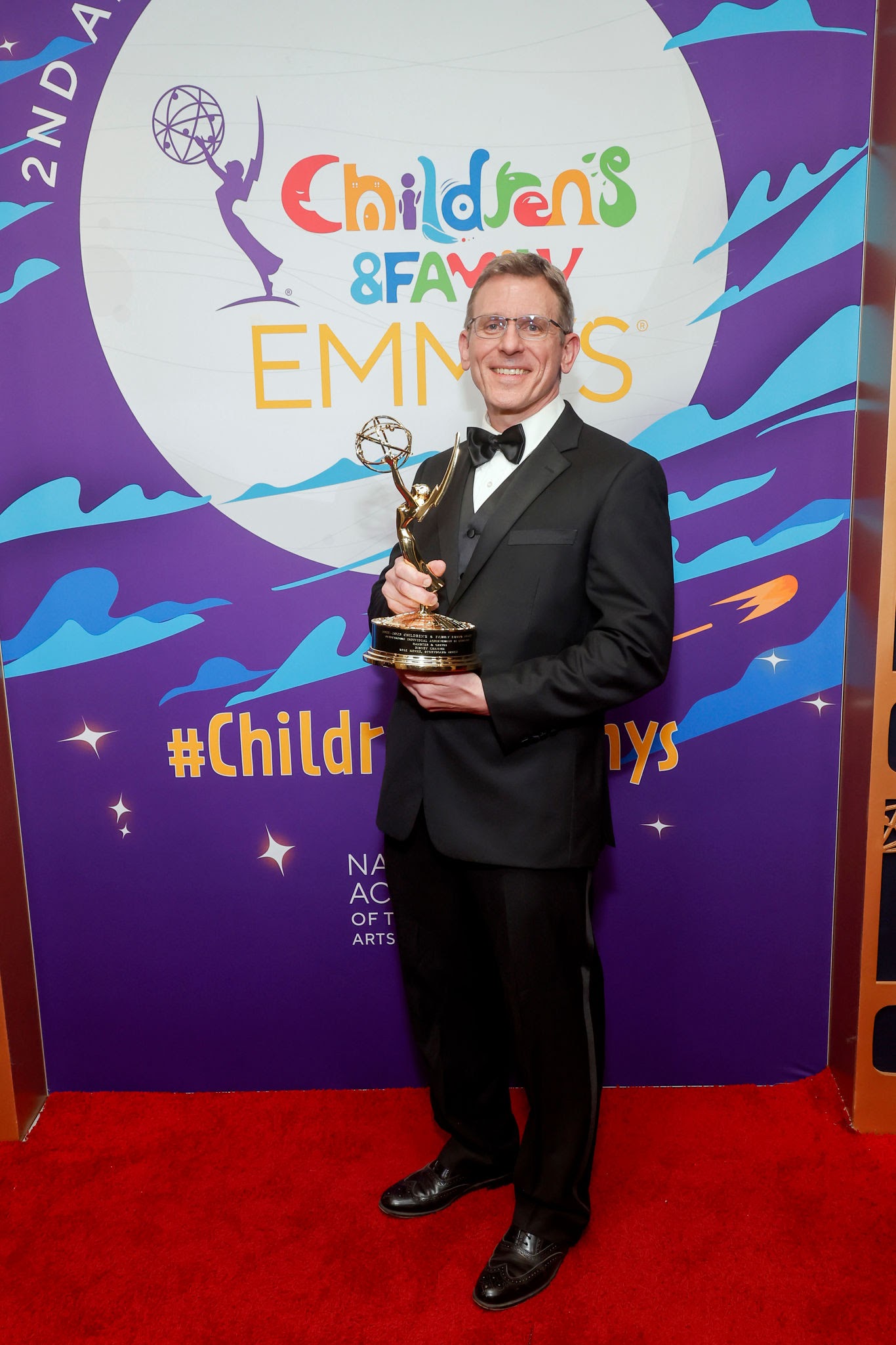 Kyle Menke in a tuxedo on the red carpet, holding his Emmy Award